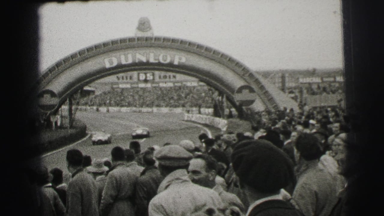 1954 Le Mans home-movie view of crowds watching race cars beneath the Dunlop arch, with foreground spectators partly blocking the track.
