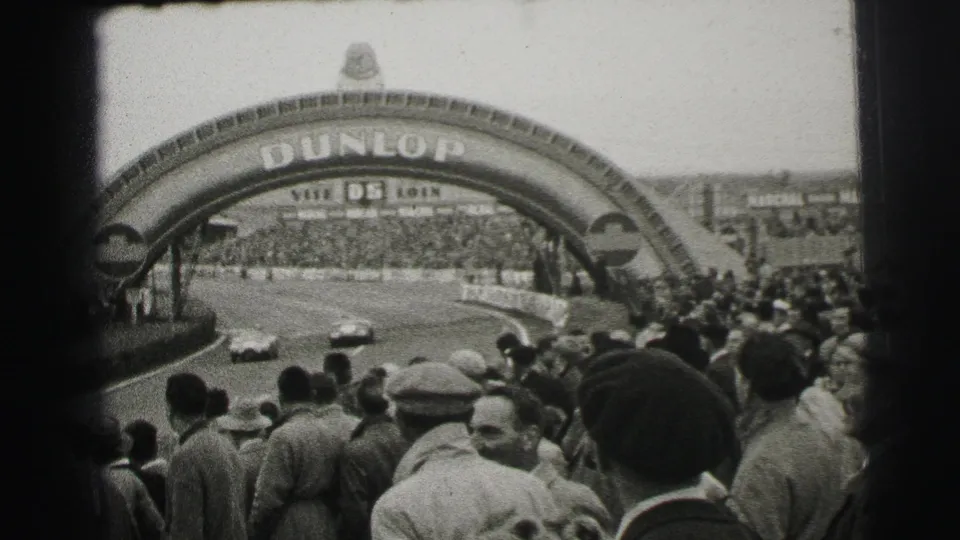 1954 Le Mans home-movie view of crowds watching race cars beneath the Dunlop arch, with foreground spectators partly blocking the track.