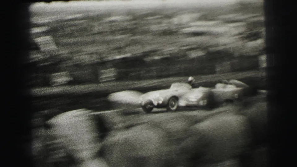 1954 Le Mans frame with strong motion blur as race cars pass close to the spectator area in black-and-white 8mm footage.