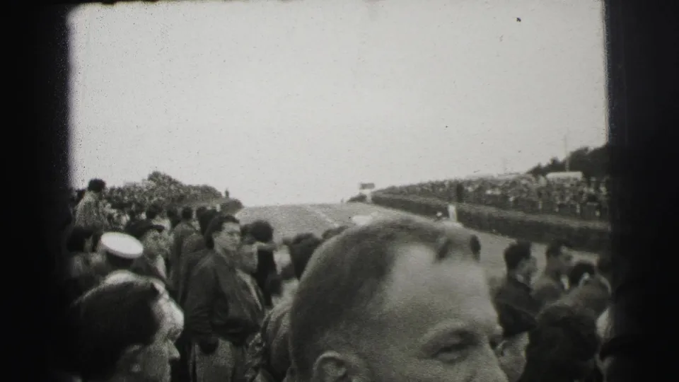 Le Mans spectator view with crowds lining both sides of the road course and a close foreground face partially filling the frame.