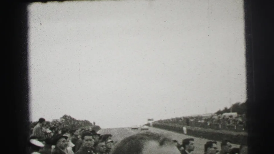 Black-and-white 1950s Le Mans crowd scene with spectators packed near the course and foreground heads partly blocking the view.