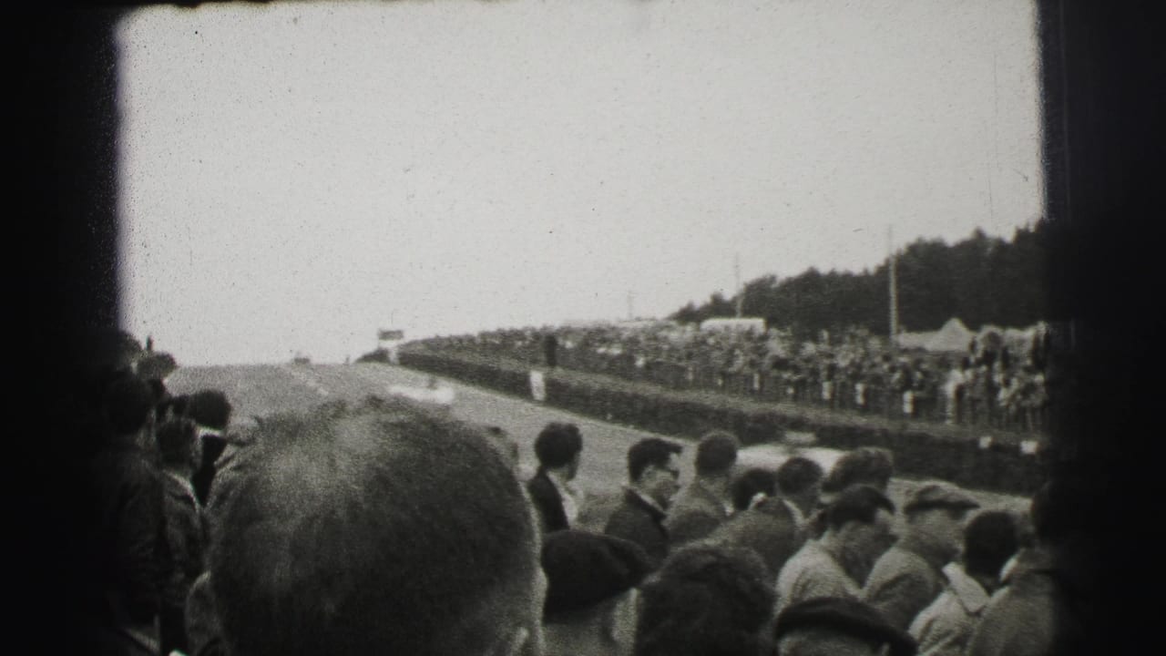 Trackside Le Mans crowd in black-and-white 8mm, with spectators in coats and caps looking toward the course from a roadside bank.