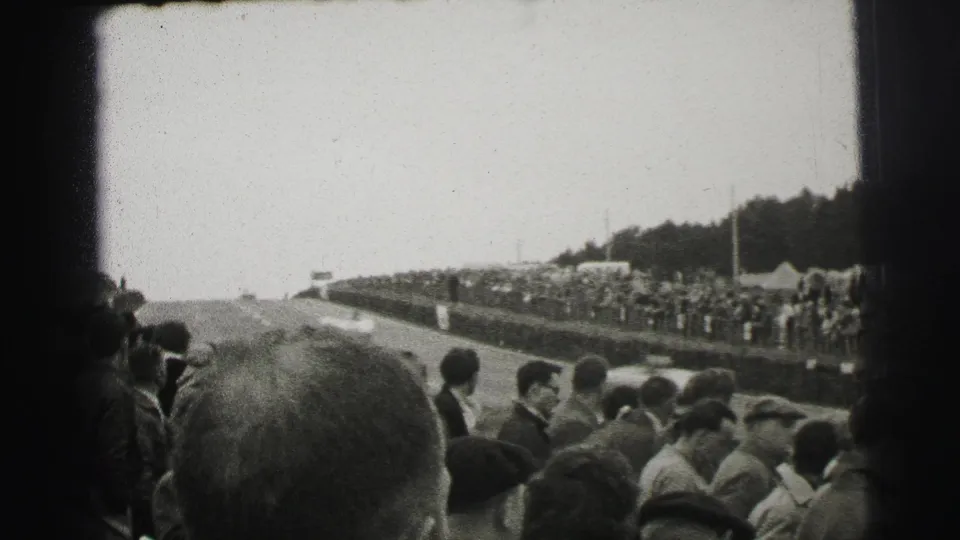 Trackside Le Mans crowd in black-and-white 8mm, with spectators in coats and caps looking toward the course from a roadside bank.