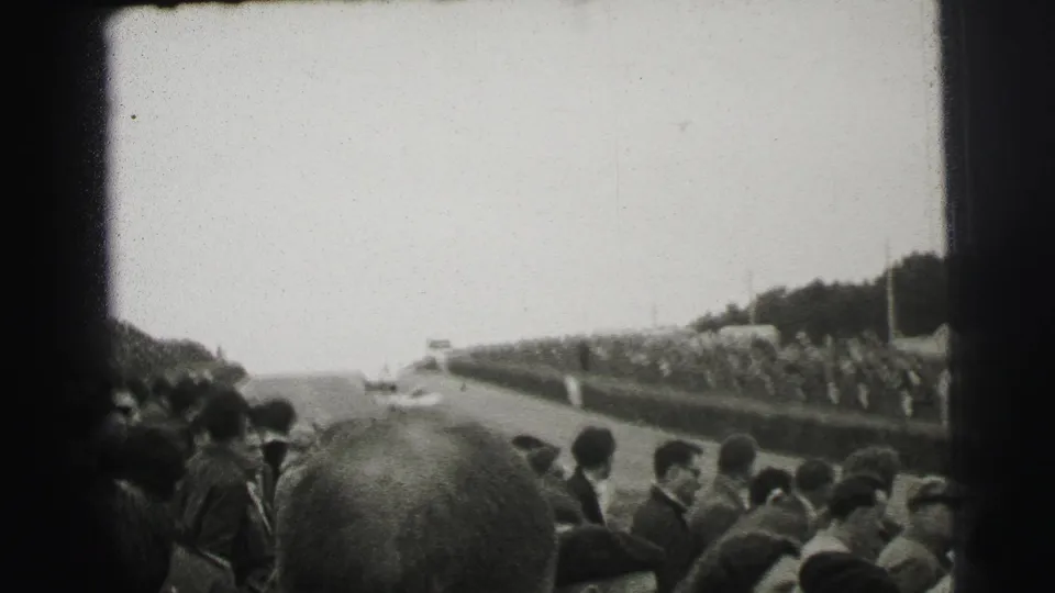 1954 Le Mans spectator view with a distant race car on the track and crowds lining the roadside barriers.