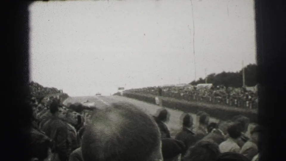 Spectators in mid-century dress line the Le Mans track as a race car appears far ahead on the road.