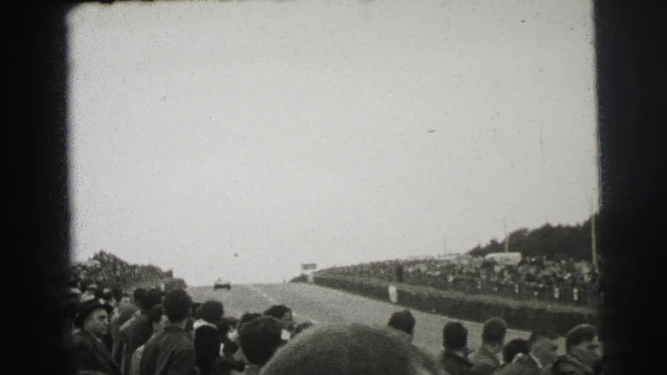 Home-movie view from the Le Mans crowd shows a race car on the course beyond foreground spectators.