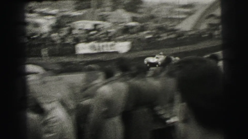At Le Mans in 1954, a race car appears blurred as it passes a dense roadside crowd filmed from a handheld 8mm spectator viewpoint.