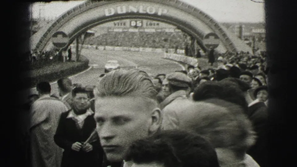 At the 1954 Le Mans setting, spectators gather beneath a large Dunlop-branded arch while people in coats watch the circuit area.