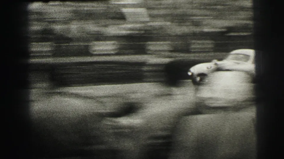 A light-colored racing car speeds past the Le Mans crowd, with strong motion blur and film edges emphasizing the handheld 8mm capture.