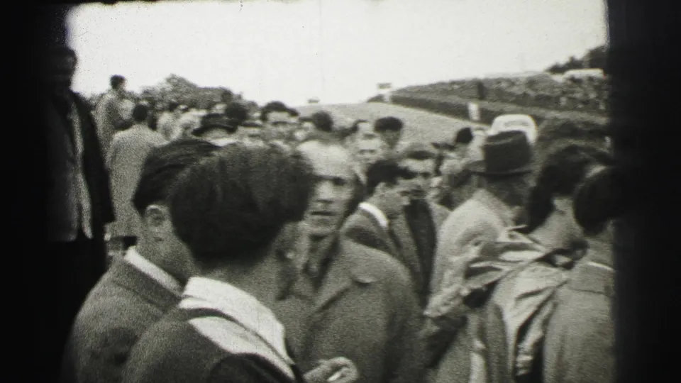Spectators at the 1954 Le Mans race stand close together near the circuit, with period coats, visible faces, and a crowded viewing area under cloudy daylight.