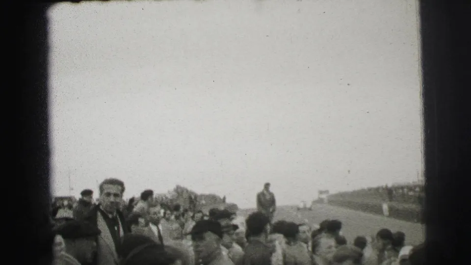 1954 Le Mans home-movie frame showing spectators along the track, with a man in a dark coat facing the camera.
