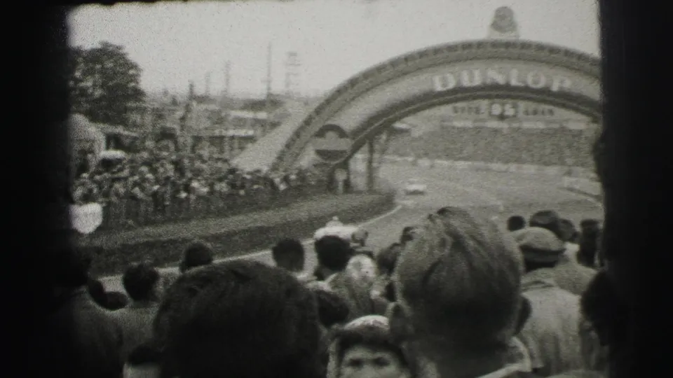 1954 Le Mans frame with spectators watching from near the Dunlop bridge as a race car approaches on the track.