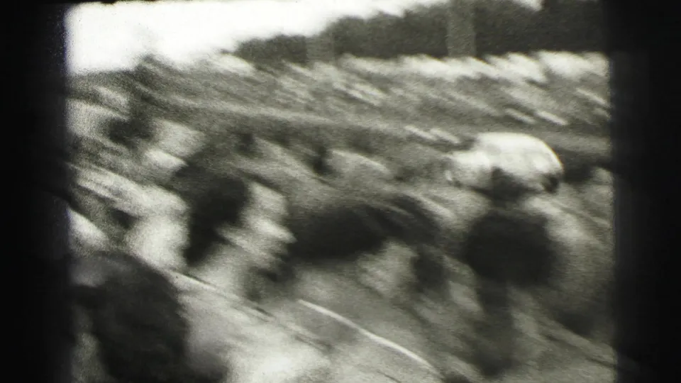 Black-and-white 8mm frame from Le Mans showing a blurred trackside crowd as a race car passes during the event.