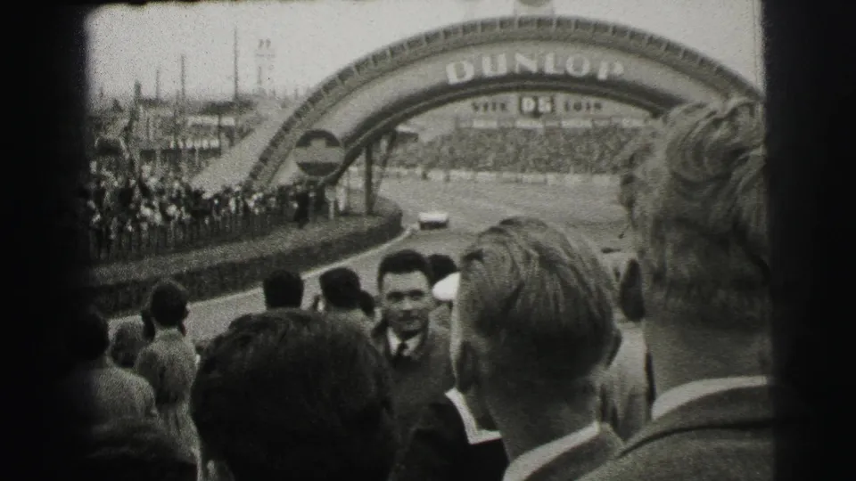 Le Mans trackside spectators face the course near the Dunlop bridge, with a race car visible on the straightaway.