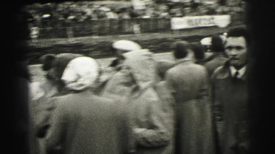 Black-and-white Le Mans crowd scene with spectators in period dress standing close to the track and grandstands behind them.
