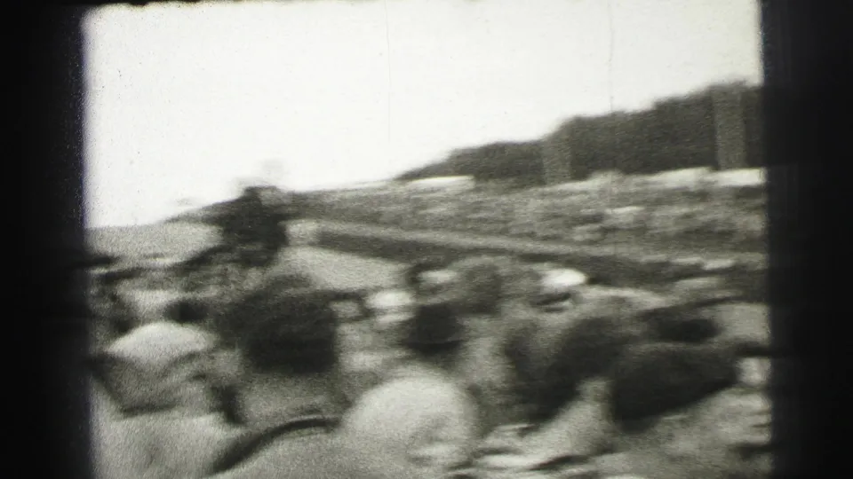 Handheld black-and-white 8mm view across Le Mans spectators toward the course and crowded grandstands.