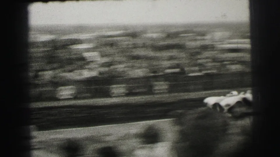 A fast-moving car blurs across the lower frame as spectators appear streaked behind it at the Le Mans course.