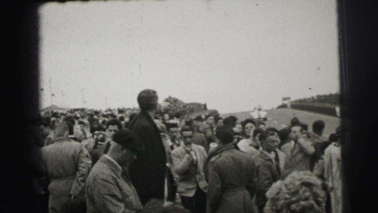 Crowd of spectators beside a race track, with men in suits and hats facing the circuit.