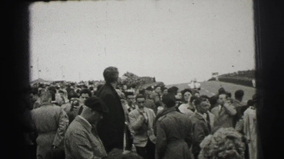 1954 Le Mans crowd scene with spectators in period clothing gathered behind barriers beside the race track.