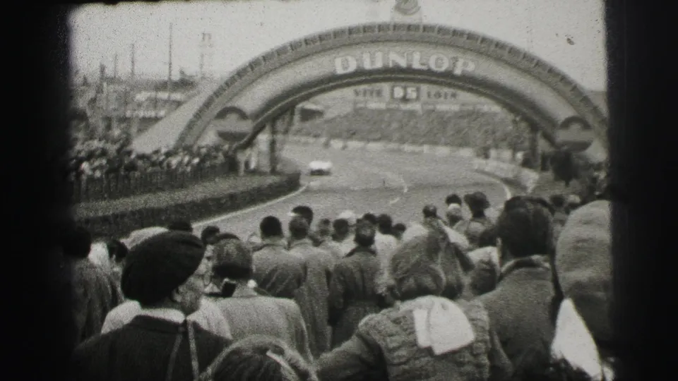 At Le Mans, spectators line the track as a light-colored race car approaches the Dunlop bridge signage.