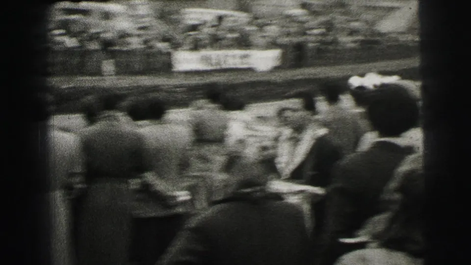 Motion-blurred Le Mans crowd view from within the spectators as a light-colored car passes on the track.