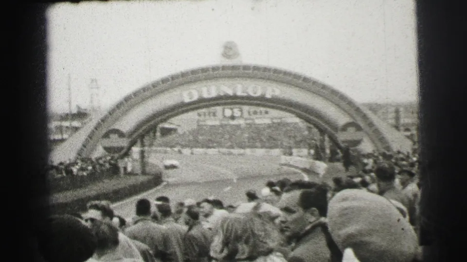 Spectators at Le Mans look toward the Dunlop arch as a light-colored race car travels along the circuit.