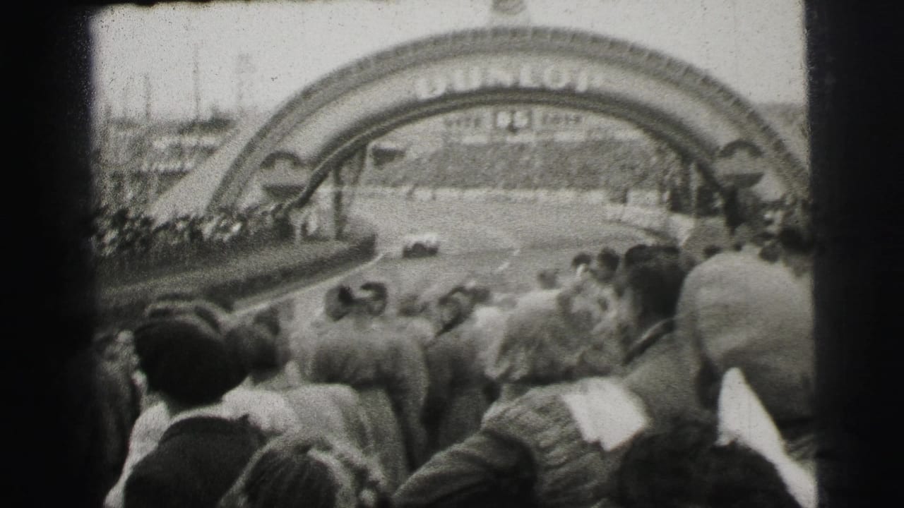 1954 Le Mans home-movie frame showing spectators facing a race car beneath the Dunlop arch.