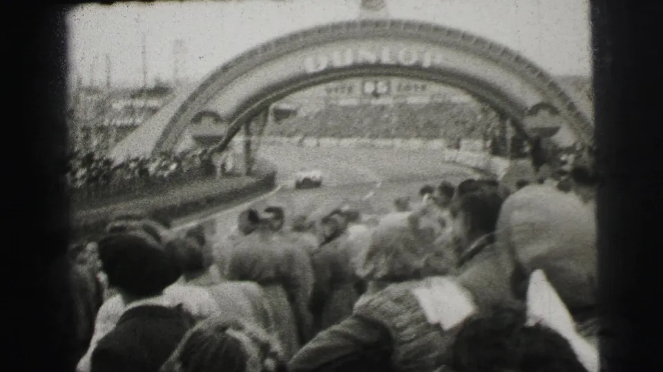 1954 Le Mans home-movie frame showing spectators facing a race car beneath the Dunlop arch.