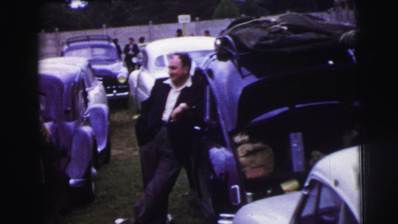 1954 Le Mans paddock or parking area with a suited man standing among rounded mid-century cars.