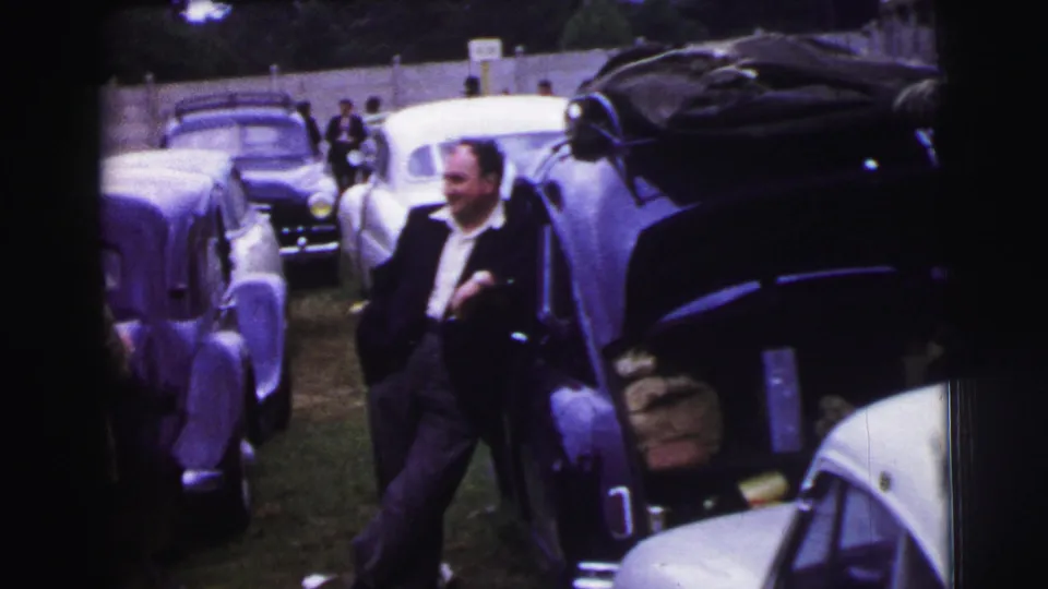 1954 Le Mans paddock or parking area with a suited man standing among rounded mid-century cars.