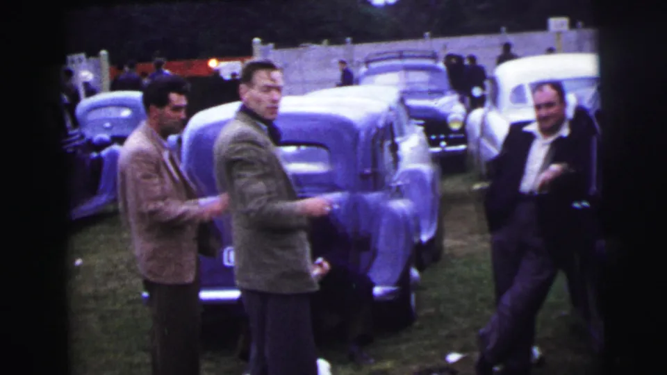 1954 Le Mans scene with men in jackets talking or looking toward the camera beside parked classic cars.