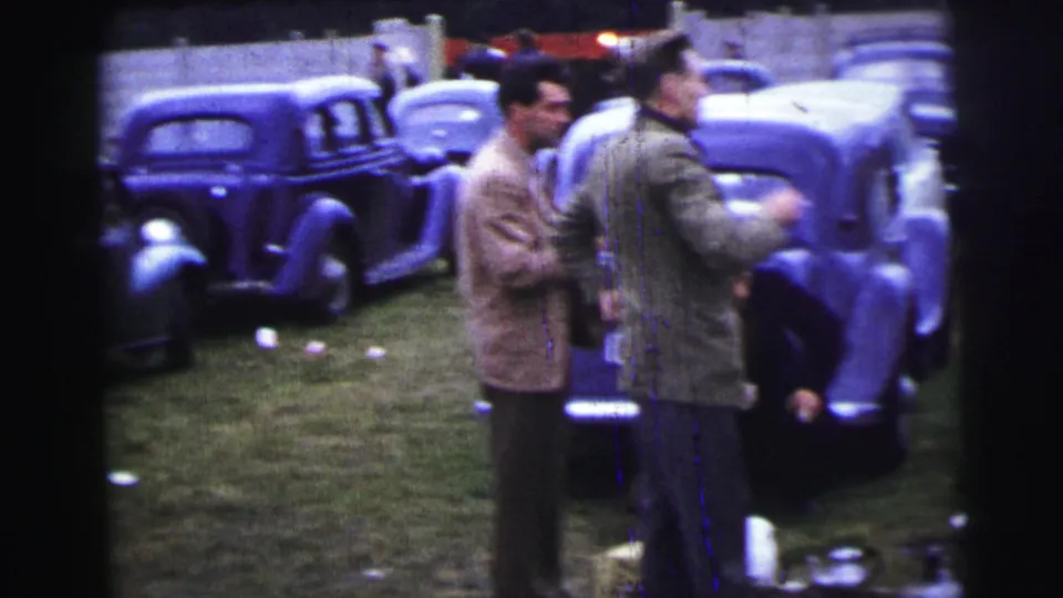 1954 Le Mans home-movie frame showing two men moving through a grassy area lined with mid-century cars.
