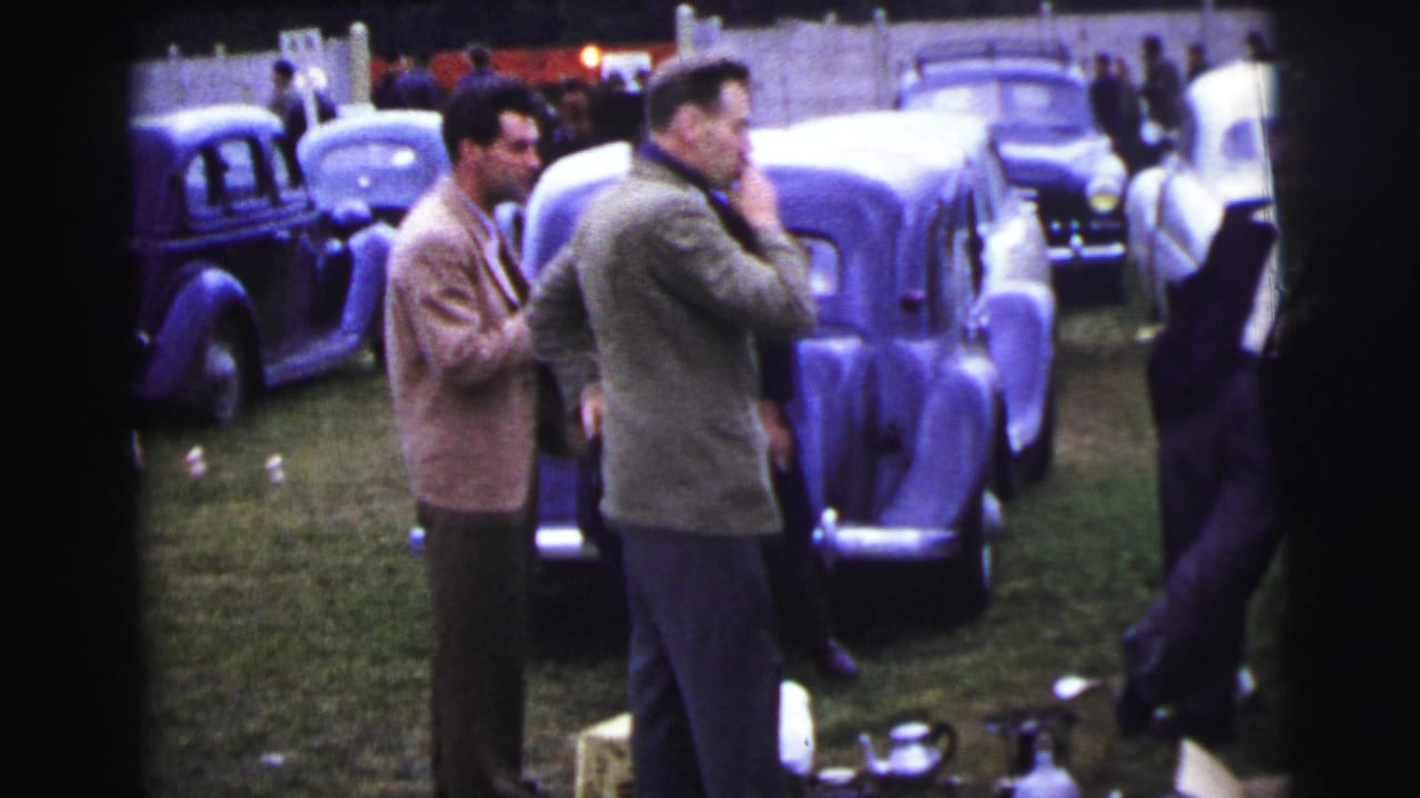 At Le Mans in 1954, suited men stand among rows of parked vintage cars on grass near a pale fence.