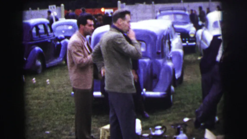At Le Mans in 1954, suited men stand among rows of parked vintage cars on grass near a pale fence.