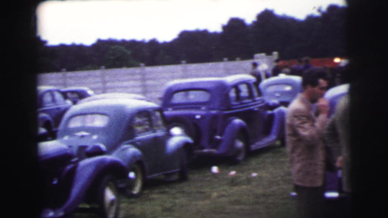 Rows of dark vintage cars sit on grass beside a pale fence.