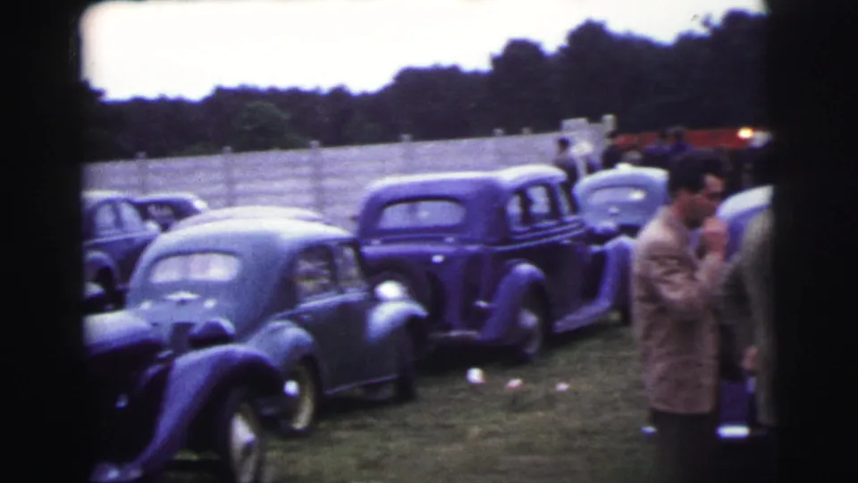 Parked vintage cars line a grassy area at the 1954 Le Mans footage location, with spectators partly visible at the edge.