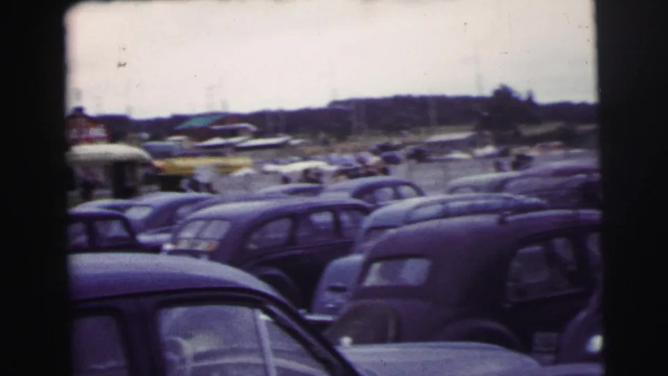 Rows of vintage cars fill the grassy Le Mans event area, with distant structures and hills beyond the parking rows.