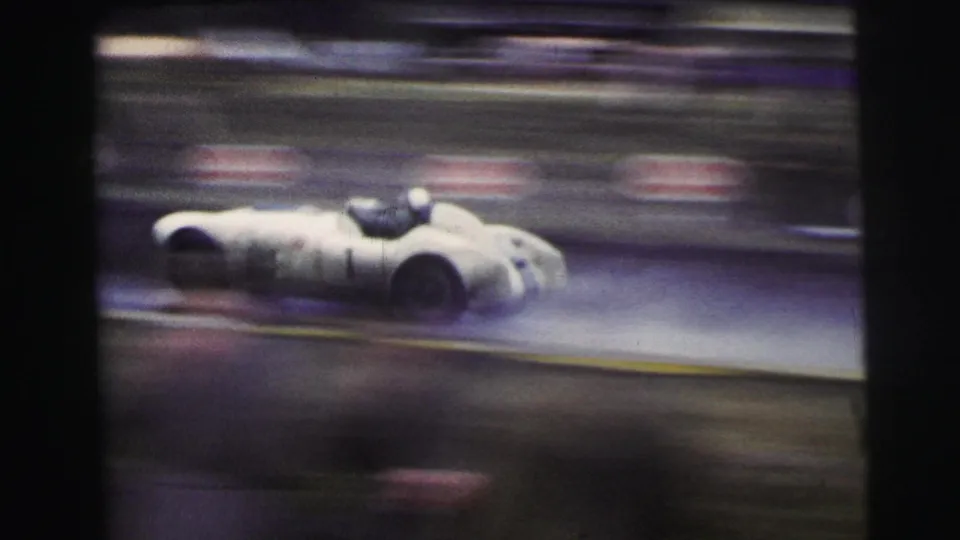 At Le Mans in 1954, a white open race car passes on a rain-dark track with strong motion blur and dark film edges.