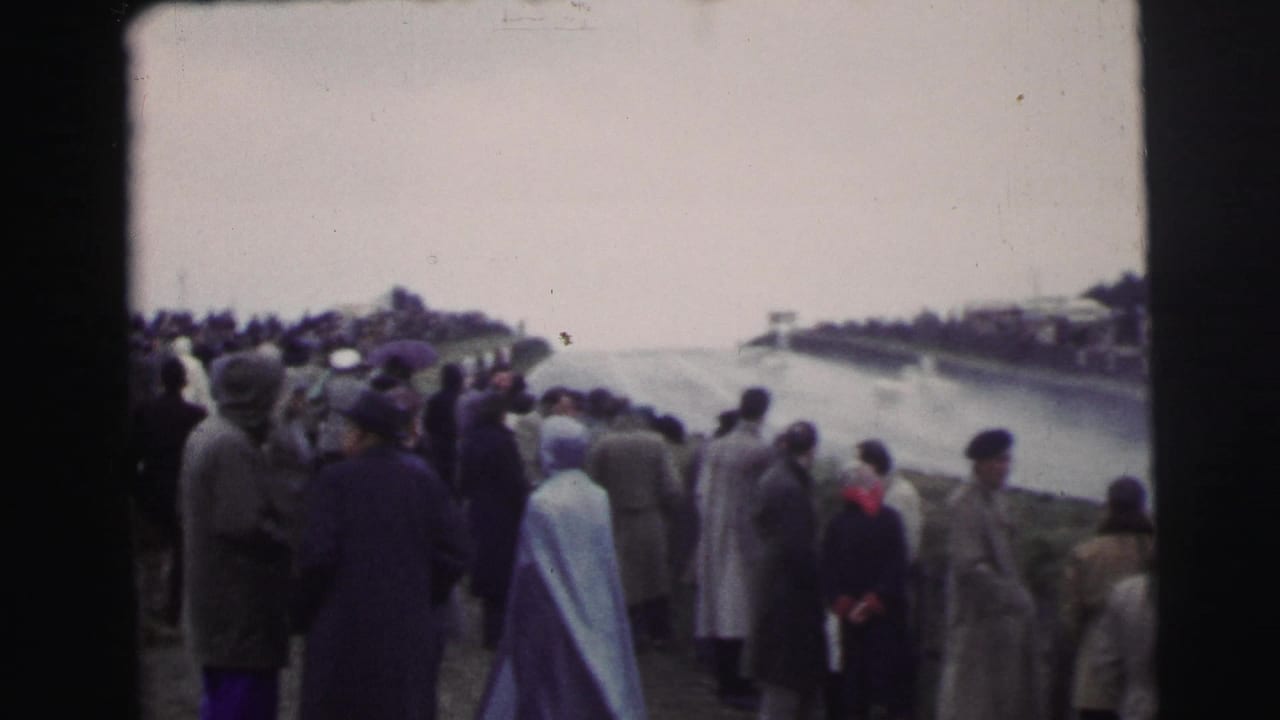Spectators in 1950s coats and hats watch from the sideline beside a wet Le Mans racing circuit under overcast light.