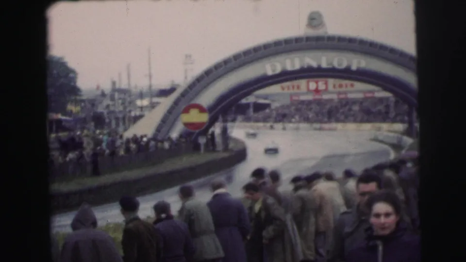 A crowd stands near the Dunlop arch at the 1954 Le Mans circuit as race cars move along the rain-wet track.