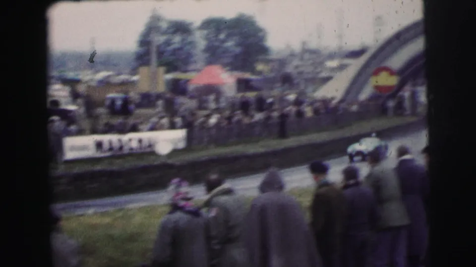 A blurred race car passes spectators near roadside signage and part of the Dunlop arch at rainy 1954 Le Mans.
