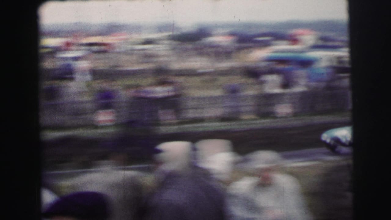 1954 Le Mans home-movie frame with blurred spectators near a track, distant tents, and parked vehicles beyond the barrier.