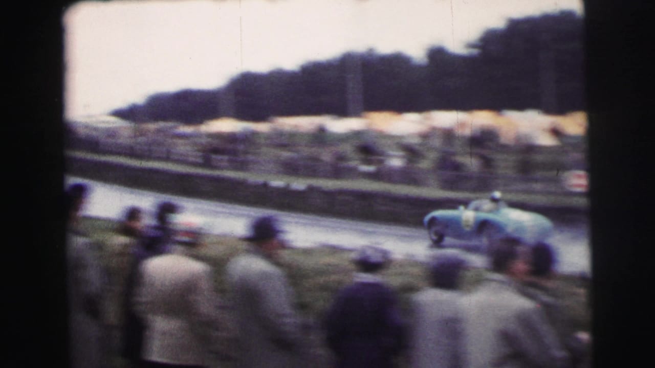 Light blue sports car passes spectators beside a tree-lined race track.