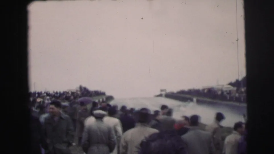 Crowded spectator view at the 1954 Le Mans race, with people in mid-century coats and hats gathered beside the track.