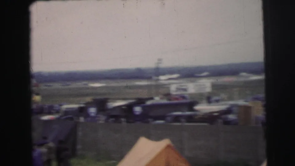 Le Mans race surroundings with parked vehicles, tents, open grass, and distant track infrastructure in a soft handheld frame.
