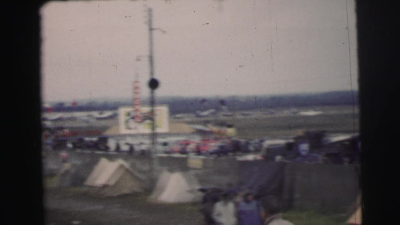 Distant race area with tents, trackside structures, and blurred spectators at Le Mans.