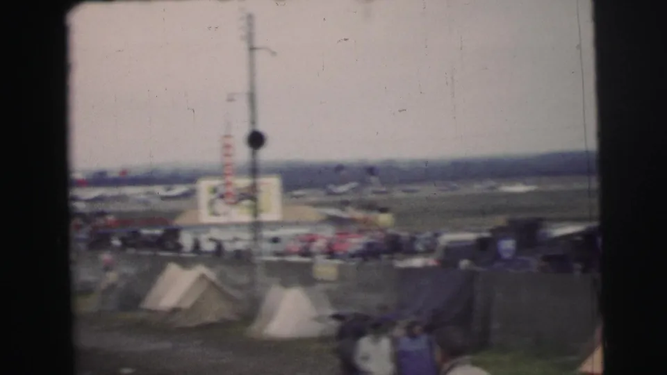 1954 Le Mans home-movie frame showing tents, trackside structures, distant cars, and spectators from a soft handheld viewpoint.