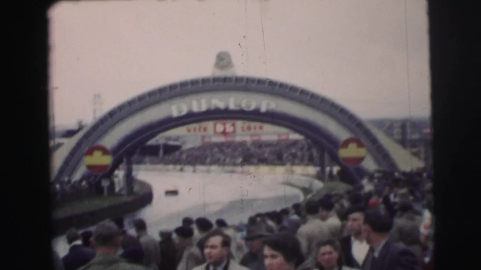 1954 Le Mans home-movie frame showing spectators along the circuit with race cars and the Dunlop arch in the background.