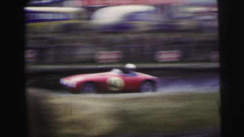 Red vintage racing car speeds past at Le Mans with spectators blurred in the background.