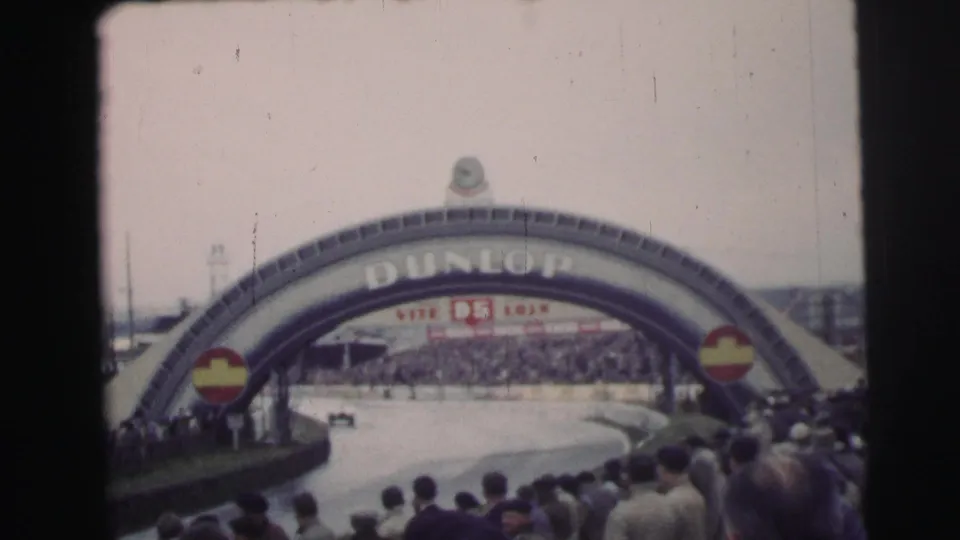 Wide 1954 Le Mans view showing the Dunlop arch, trackside crowd, and race course in soft 8mm color.